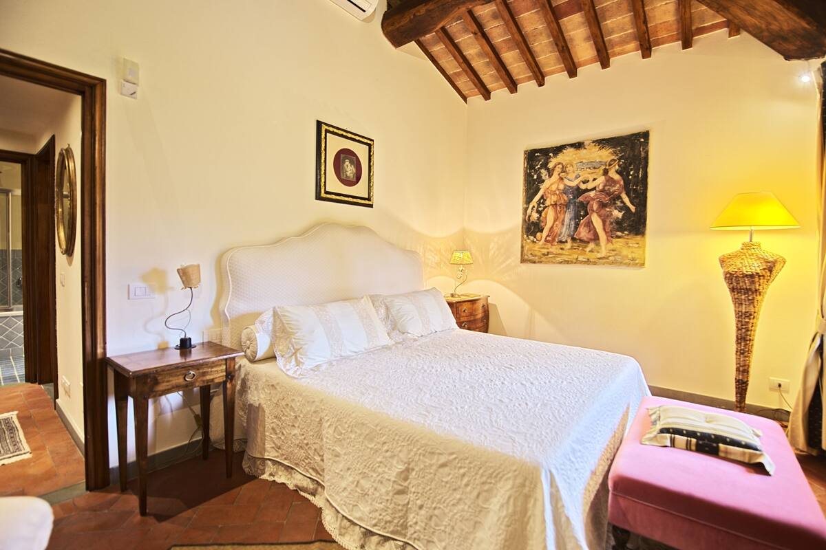 A bedroom with a large bed, a nightstand, a lamp, a painting, and a small table.