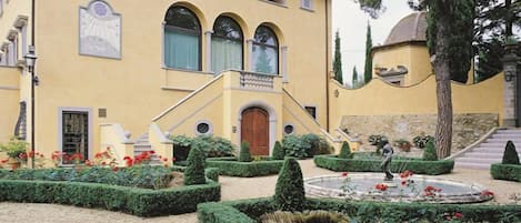 A yellow building with a central staircase, a garden with trimmed hedges, and a fountain.