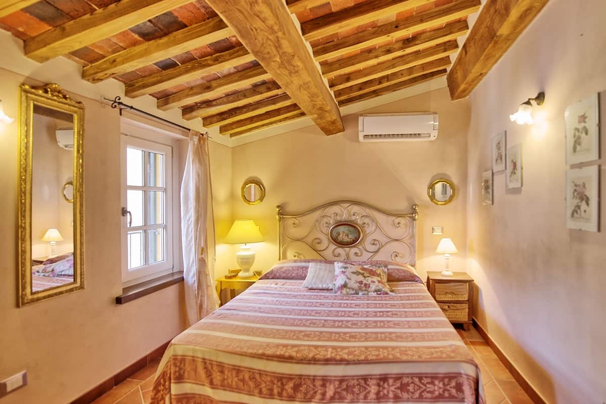 A bedroom with a wooden ceiling, a bed with a patterned bedspread, two bedside lamps, a mirror, and a window with curtains.