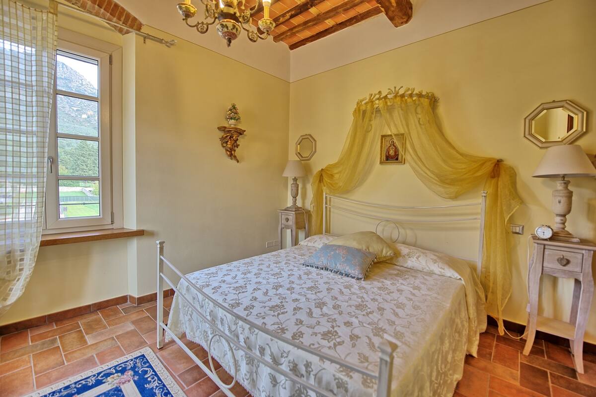 A bedroom with a bed, bedside tables, a window with curtains, a chandelier, and a mirror on the wall.
