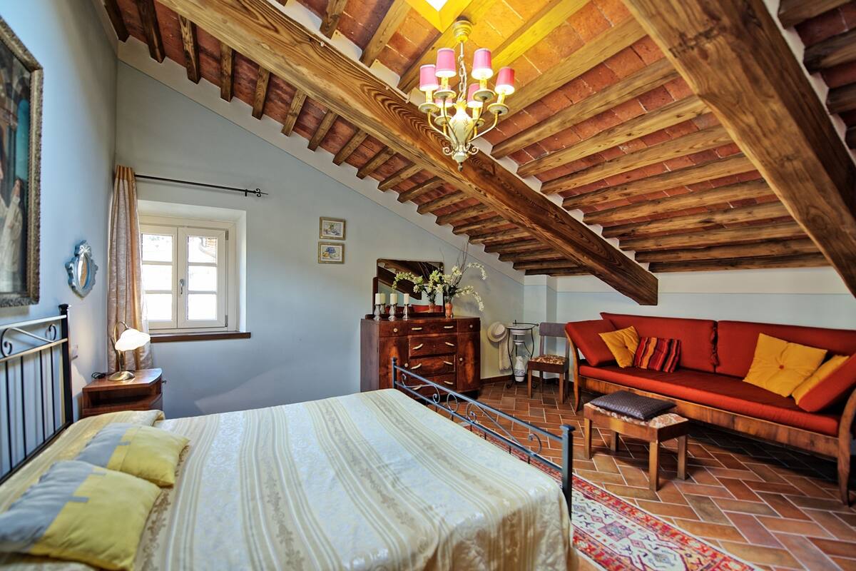 A bedroom with a bed, a red sofa, a wooden dresser, a chandelier, and a window with curtains.