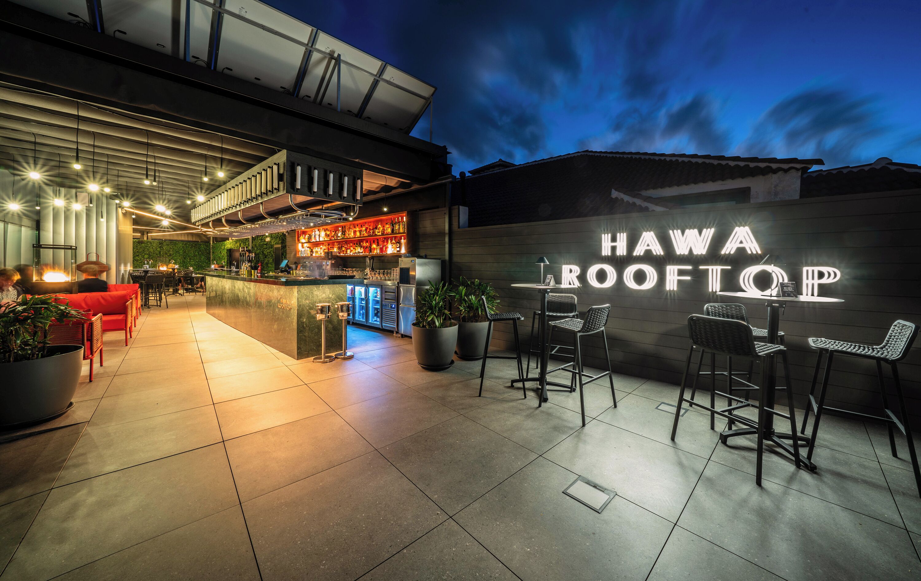 Uma área de bar ao ar livre com assentos, um letreiro de neon que diz "HAWA BOOTTOP" e um design arquitetônico moderno.