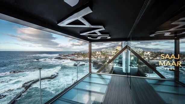 Un intérieur moderne avec un plancher de verre donnant sur l’océan, un plafond au design géométrique et le texte « SAAJ MAAR » bien visible.