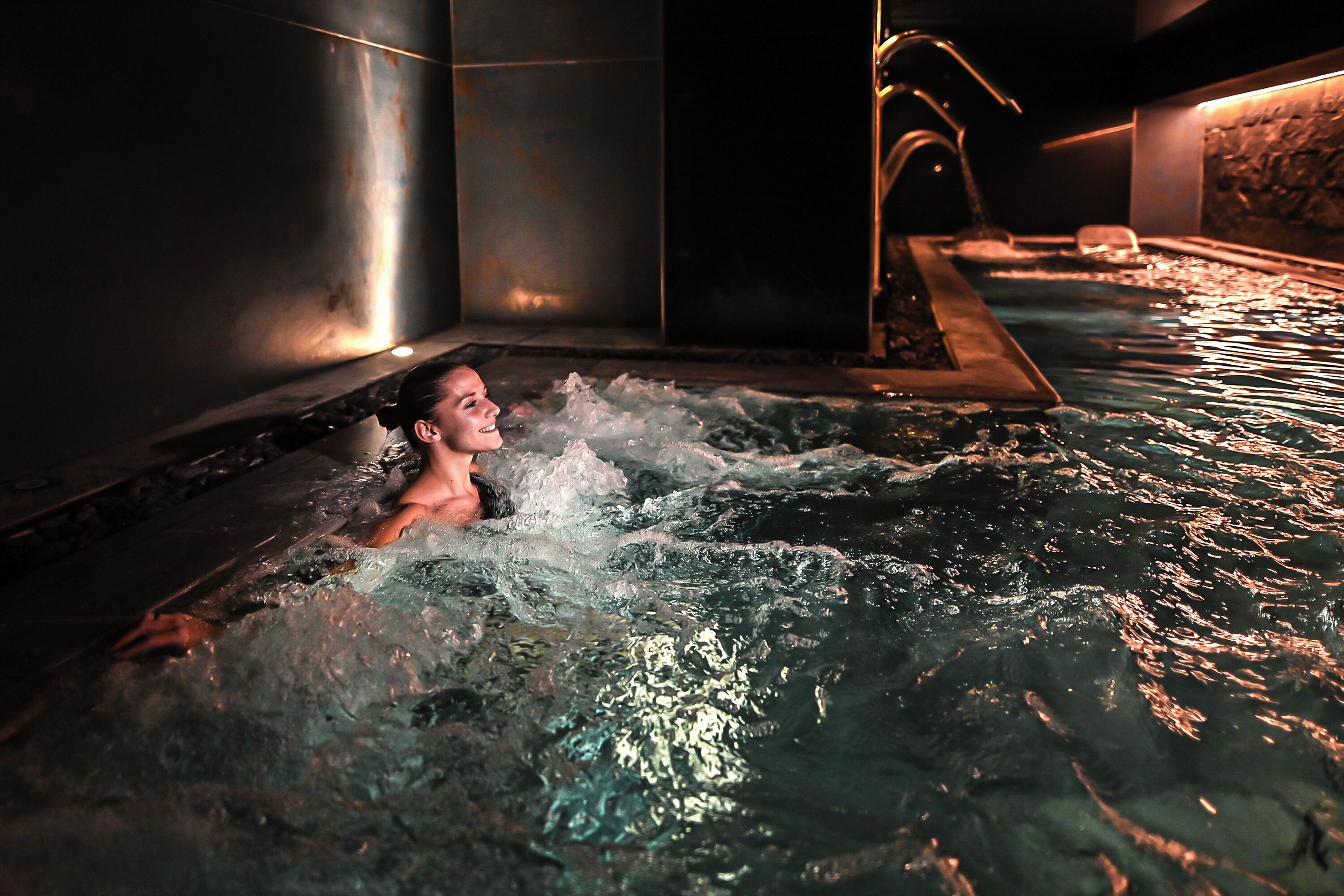 Eine Person in einer Whirlwanne mit Wasserdüsen, umgeben von Steinsäulen und gedämpftem Licht.
