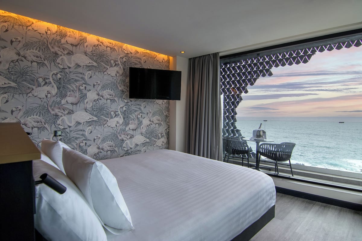 Habitación de hotel con cama, televisor montado en la pared, ventana con vistas al mar y balcón con mesa y sillas.