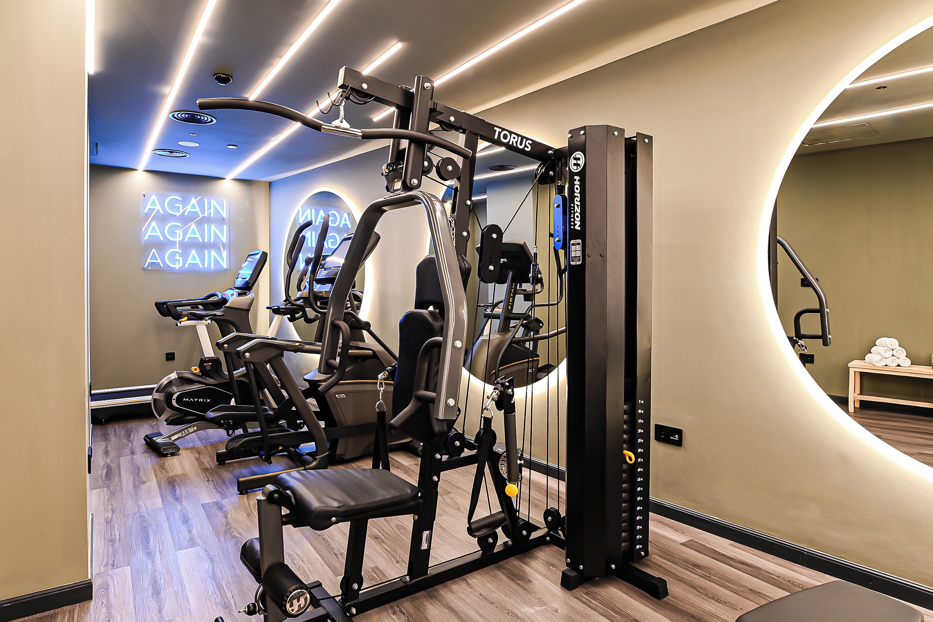 Ein modernes Fitnessstudio mit verschiedenen Trainingsgeräten, einem an der Wand montierten Bildschirm, der "AGAIN AGAIN" anzeigt, und einer Bank.