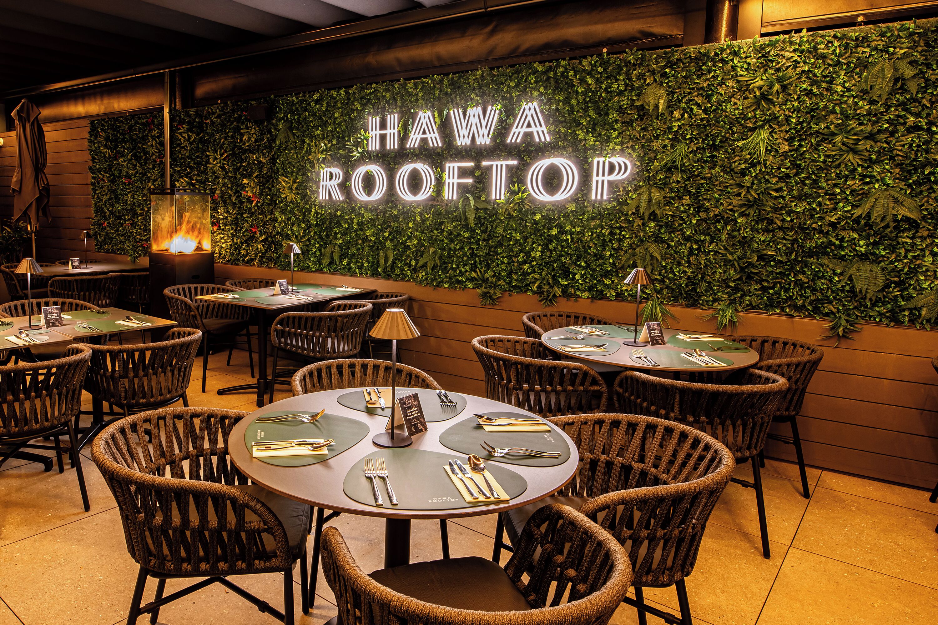 L’intérieur d’un restaurant avec des tables rondes, des chaises en osier et un panneau néon sur lequel on peut lire « HAWA ROOFTOP ».