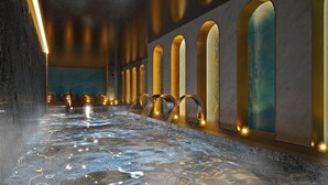 Un espace piscine intérieure luxueux, doté d’un long bassin étroit, de plusieurs éléments décoratifs aquatiques et d’une rangée d’arches le long des murs.