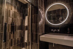 Un baño moderno con un espejo grande, paredes de azulejos oscuros y un lavamanos blanco.