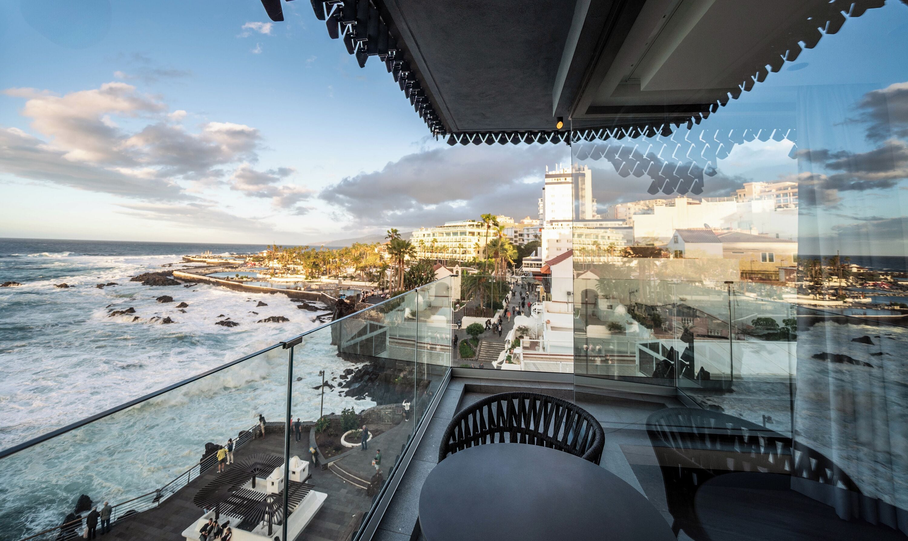 Ein Balkon mit Glasgeländer, der auf eine Küstenstadt mit Blick auf das Meer, eine Promenade und Gebäude blickt.