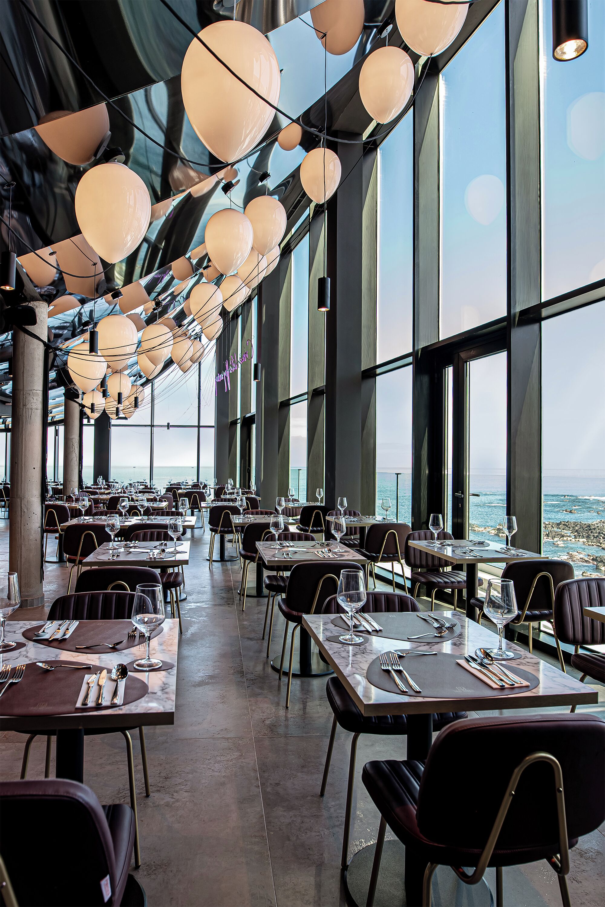 A spacious restaurant with high ceilings, large windows, and a view of the sea. Tables are set with fine dining ware.