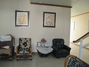 A living room with two framed floral paintings, a wicker chair, a black leather chair, a small table with a lace tablecloth, and a 'No Smoking' sign.