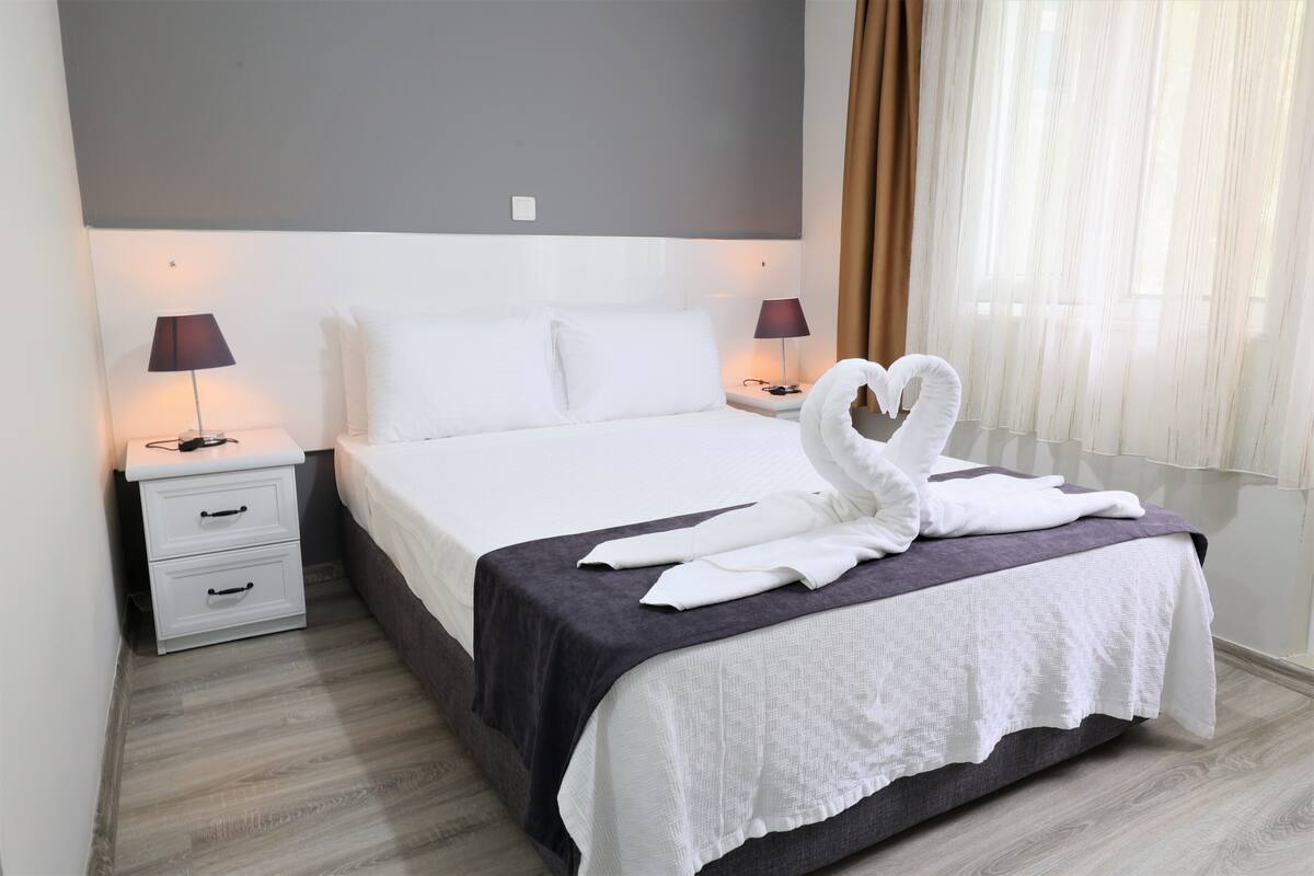 Cama bem arrumada com lençóis brancos, colcha escura e arranjo decorativo de toalhas ao lado. Há duas mesas de cabeceira com abajures, um móvel com gavetas branco e uma janela com cortinas leves.