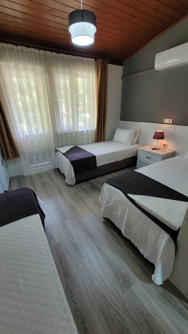 A hotel room with two beds, a nightstand with a lamp, a window with curtains, and an air conditioning unit on the wall.