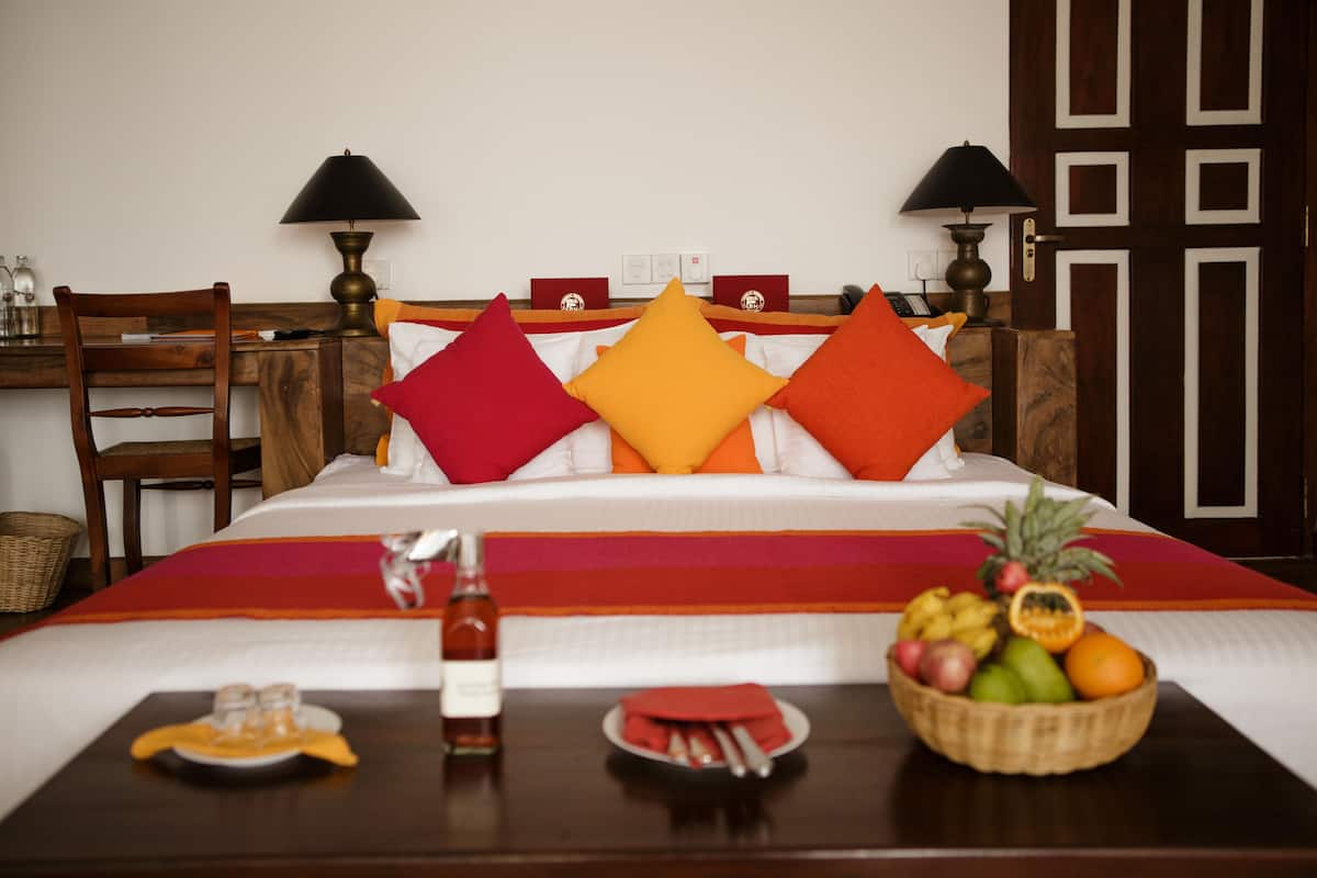 A neatly made bed with a red and white bedspread, two lamps, a wooden chair, a small table with a fruit basket, and a bottle of water.