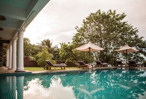Una piscina con sillas de descanso y sombrillas, rodeada de árboles y un muro de ladrillo.