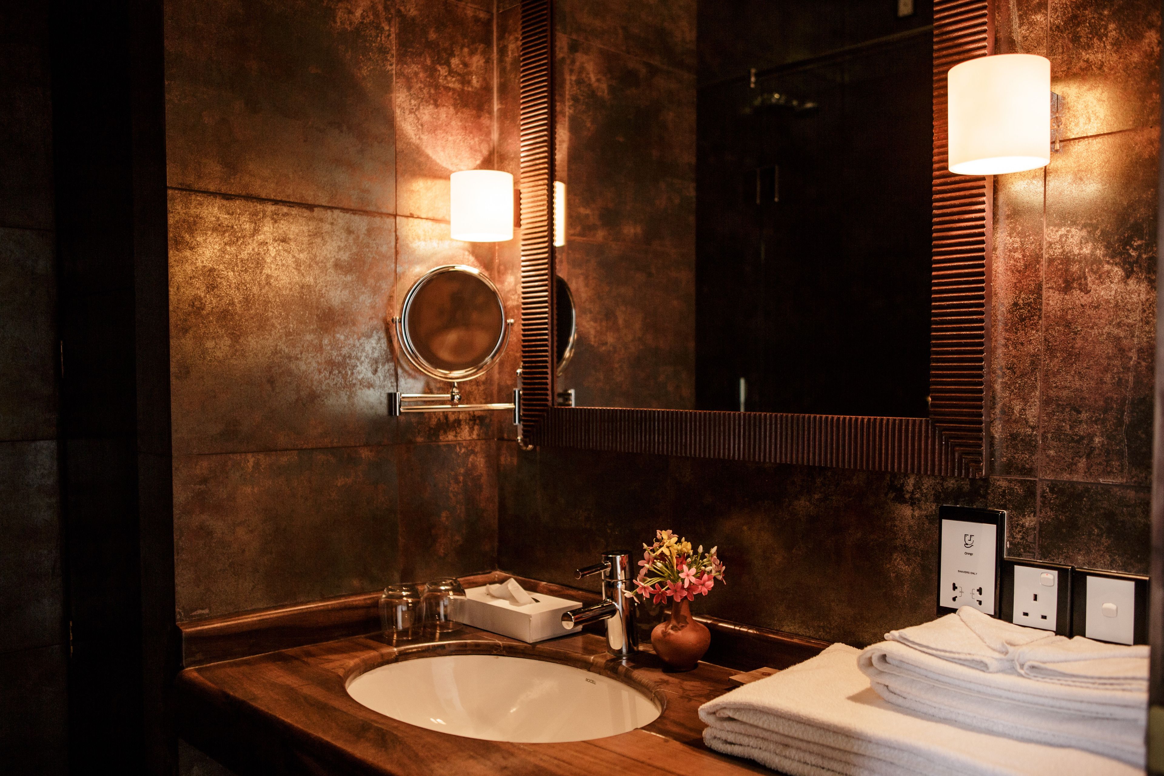 A bathroom with a wooden vanity, a round mirror, a sink, a vase with flowers, and a stack of towels.
