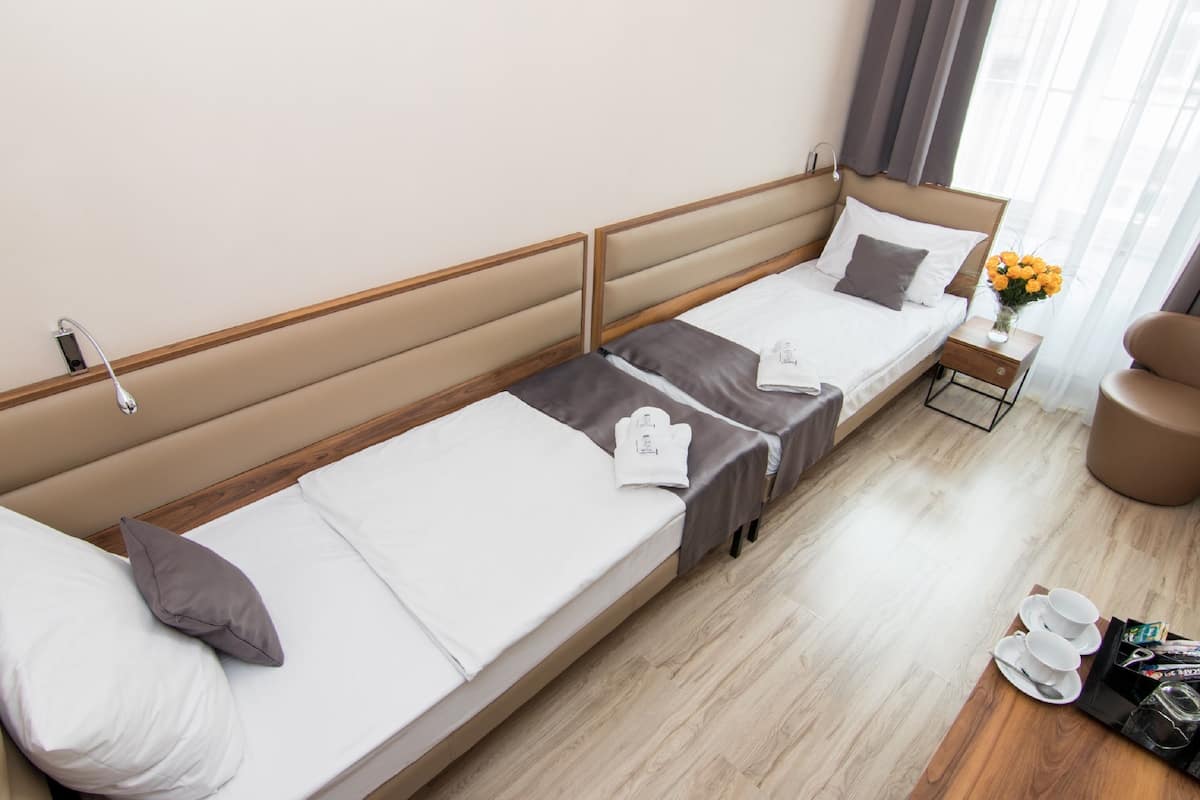 A hotel room with two single beds, a small bedside table with a vase of flowers, a chair, and a tray with cups and a bag.