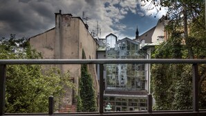 A view through a window with a metal railing, featuring a building with a glass facade and a traditional structure with a sloped roof in the background. - G12 Aparthotel (Kraków)