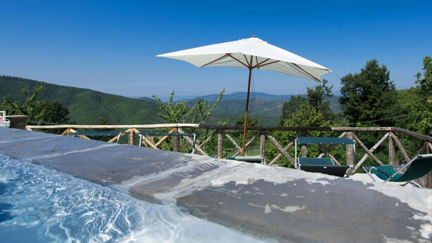A swimming pool with a wooden deck and lounge chairs, surrounded by a wooden fence and trees, with a view of distant hills. - Villa Margherita Tre (Cortona)