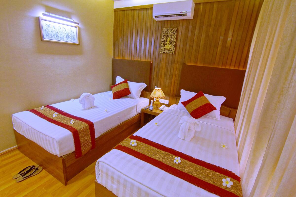 Ein Hotelzimmer mit zwei Einzelbetten, jeweils mit roter und weißer Bettdecke, einem Nachttisch mit Lampe und einer Klimaanlage an der Wand.