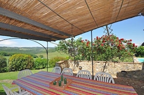 An outdoor patio with a table and chairs, a thatched roof, and a view of a garden with red flowers. - Villa Giulia (San Gimignano)