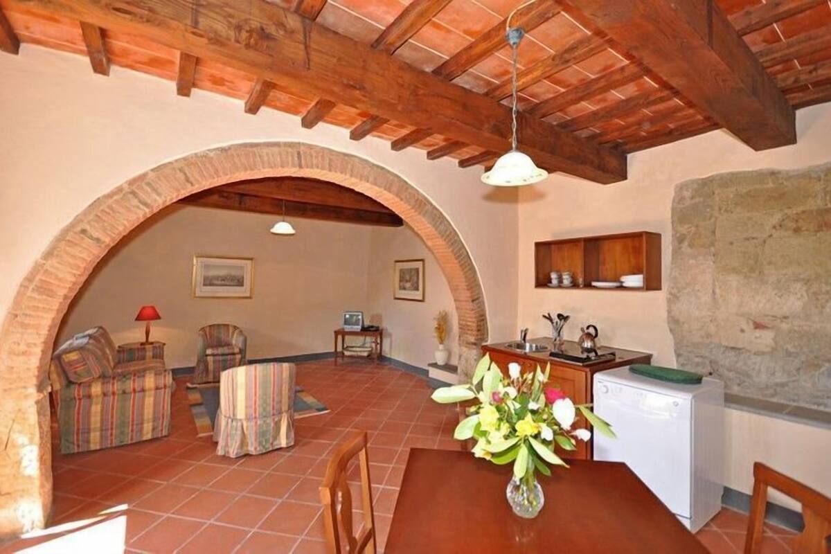 A rustic interior with exposed wooden beams, a dining area with a table and chairs, a kitchen with a sink and a small refrigerator, and a living area with a sofa and armchairs.