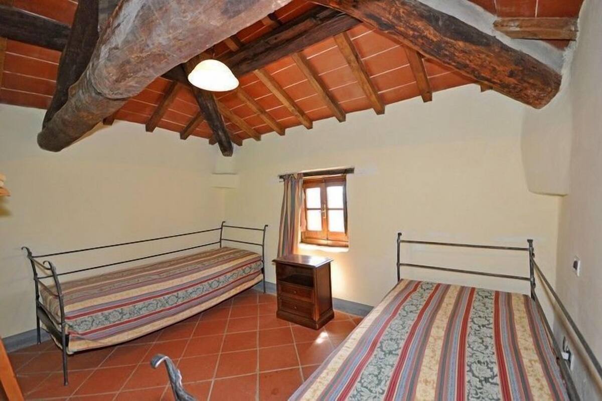 A small, rustic room with two single beds, a wooden bench, a small table, and a window with curtains.
