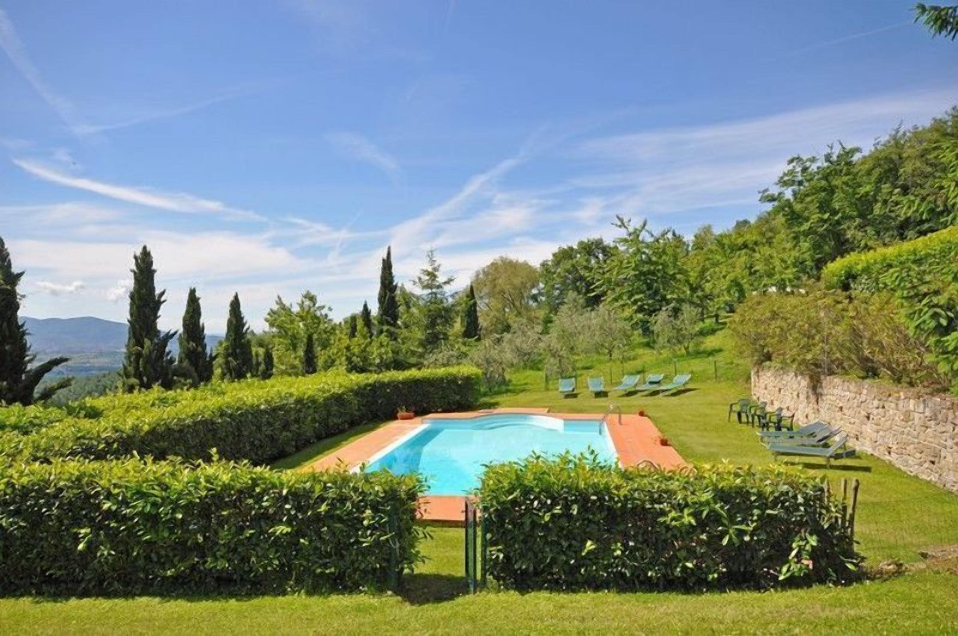 Una piscina rodeada de setos cuidados, con un muro de piedra y sillas de descanso a un lado.
