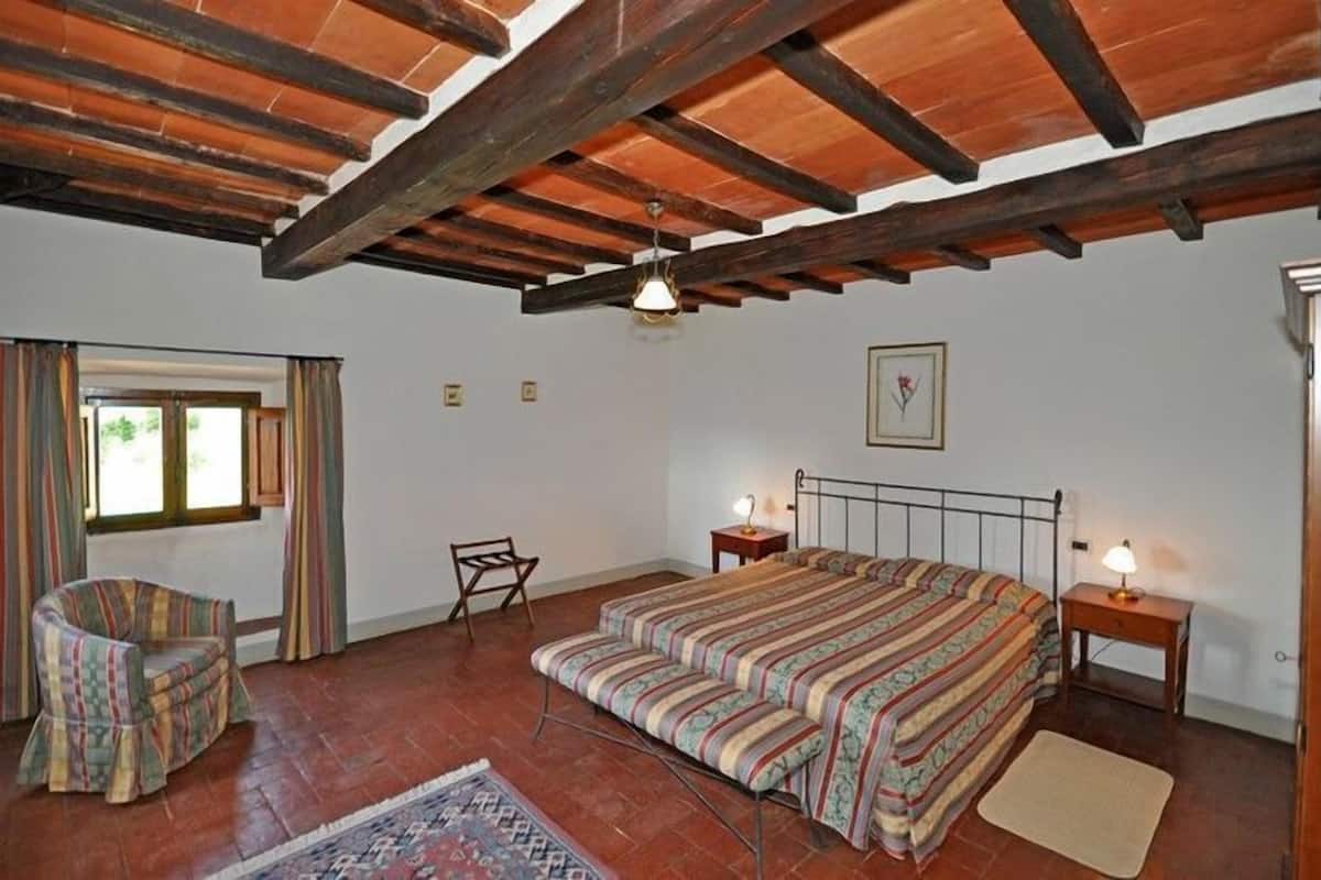 A bedroom with a wooden ceiling, a bed with striped bedding, a chair, a bedside table with a lamp, and a window with curtains.