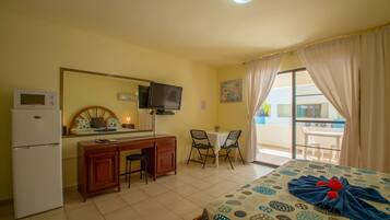Habitación de hotel con una cama, un escritorio con silla, refrigerador, televisor y balcón con mesa y sillas.