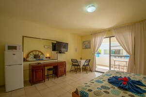 Habitación de hotel con una cama, un escritorio con silla, refrigerador, televisor y balcón con mesa y sillas.