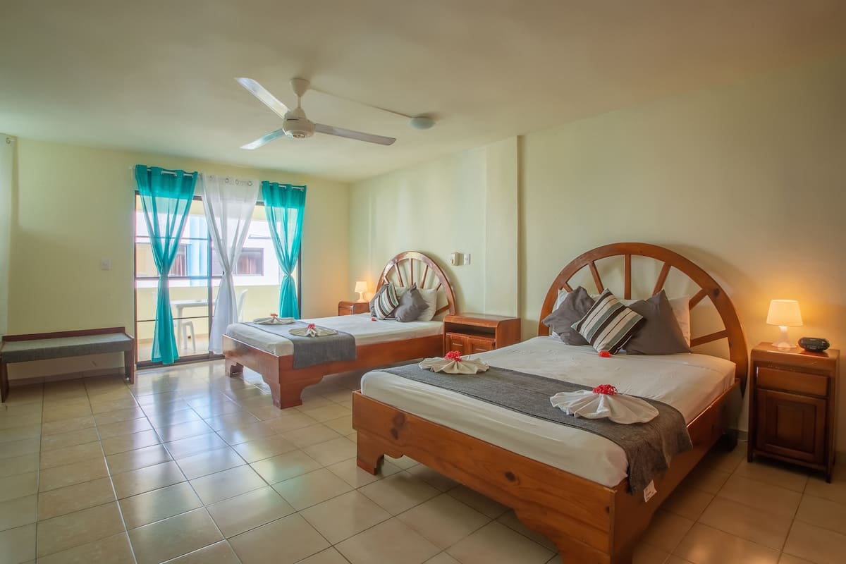 A hotel room with two wooden beds, a bench, a ceiling fan, and a balcony with curtains.