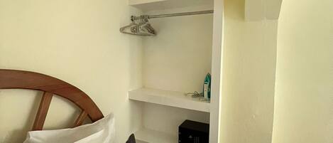 A hotel room with a bed, pillows, a bedside table, a shelf with a few items, and a wardrobe.