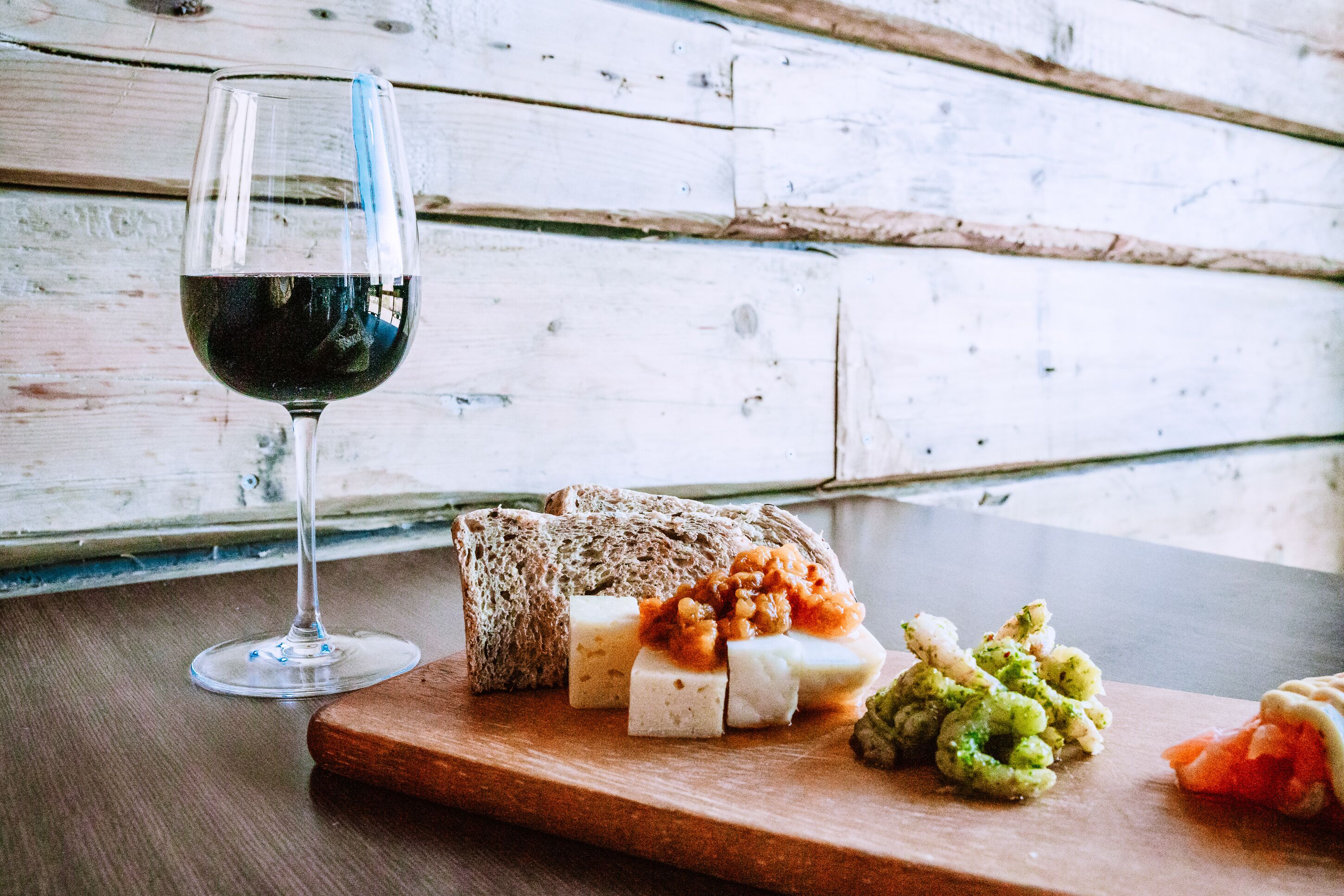A wooden table with a glass of red wine, a loaf of bread, and various cheeses and pickles.
