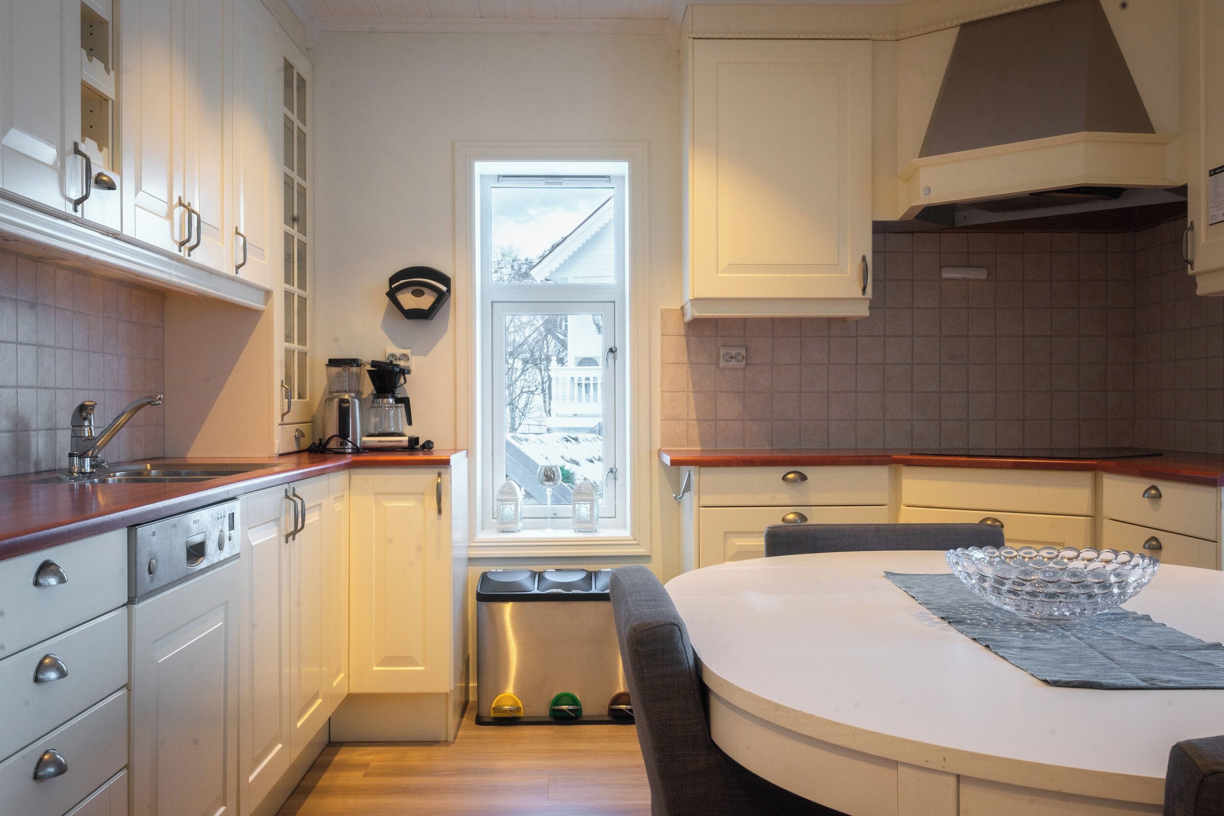 Una cocina compacta con armarios blancos, encimera de madera, lavavajillas integrado y ventana con vista a una casa.