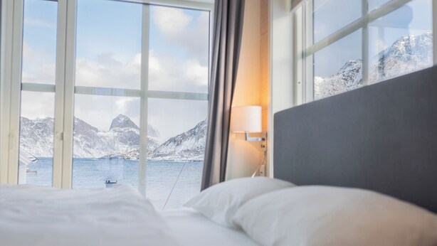 Habitación de hotel con cama, mesita de noche con lámpara y un amplio ventanal que ofrece vistas a montañas nevadas y una masa de agua.