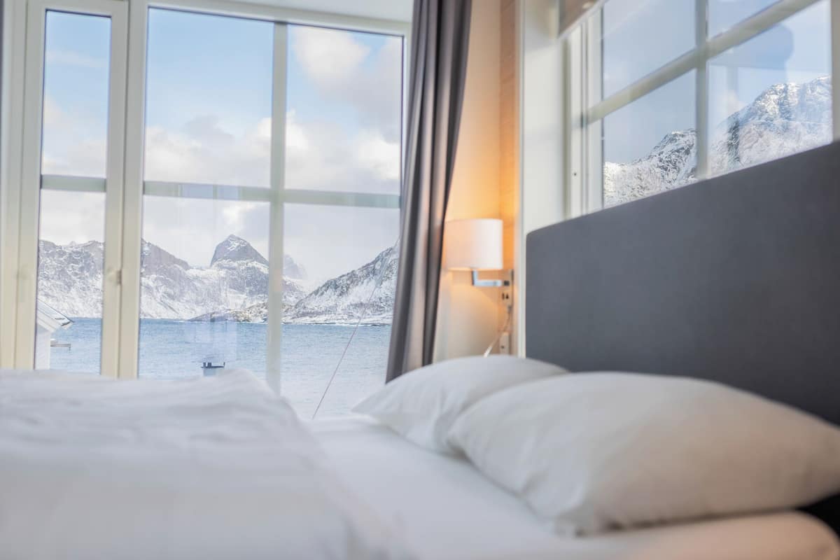 Une chambre d’hôtel avec un lit, une table de chevet avec une lampe et une grande fenêtre donnant sur un paysage de montagnes enneigées et un plan d’eau.