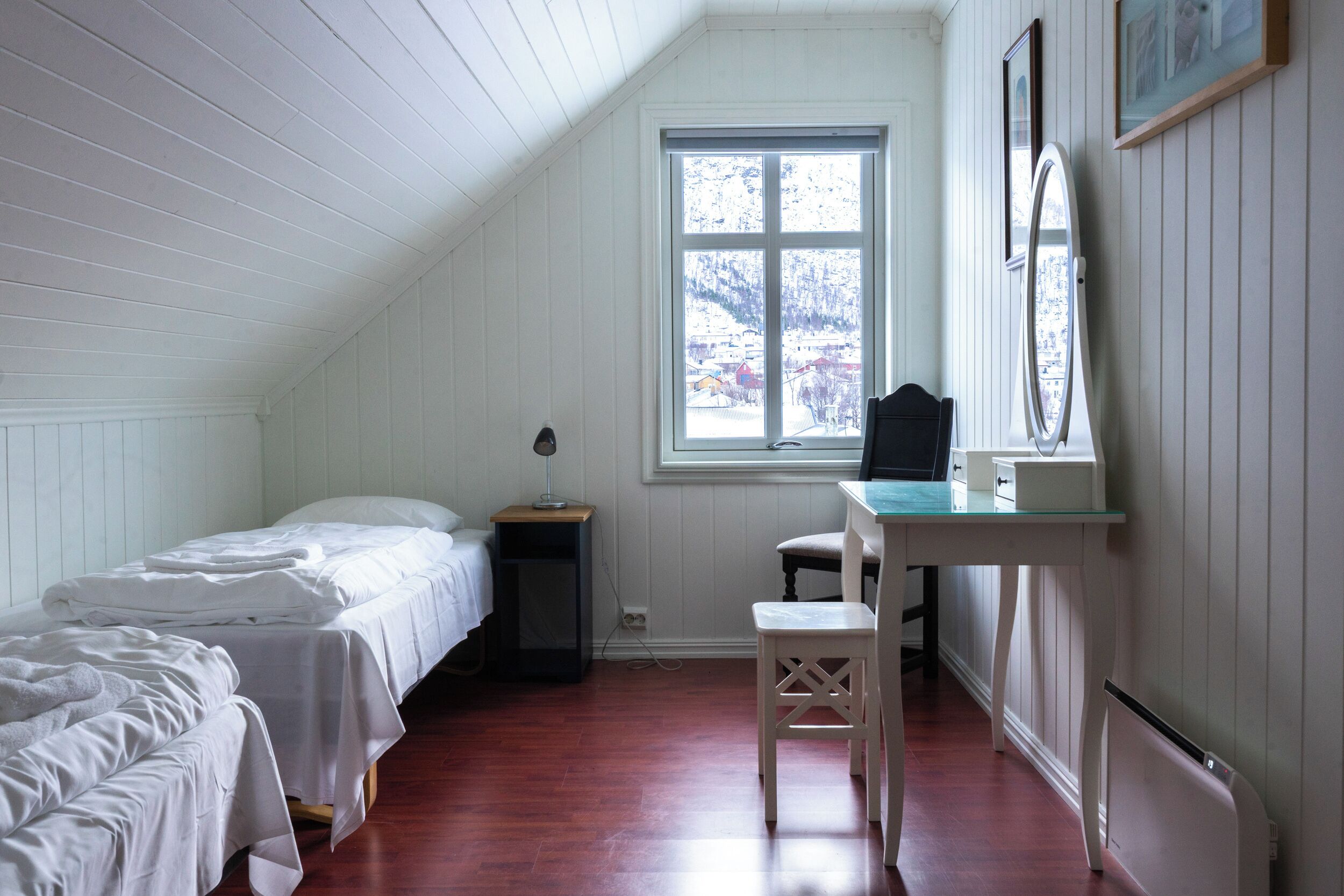 Una habitación pequeña con paneles blancos, una cama individual, un escritorio con silla y una ventana que ofrece vistas a un paisaje nevado.