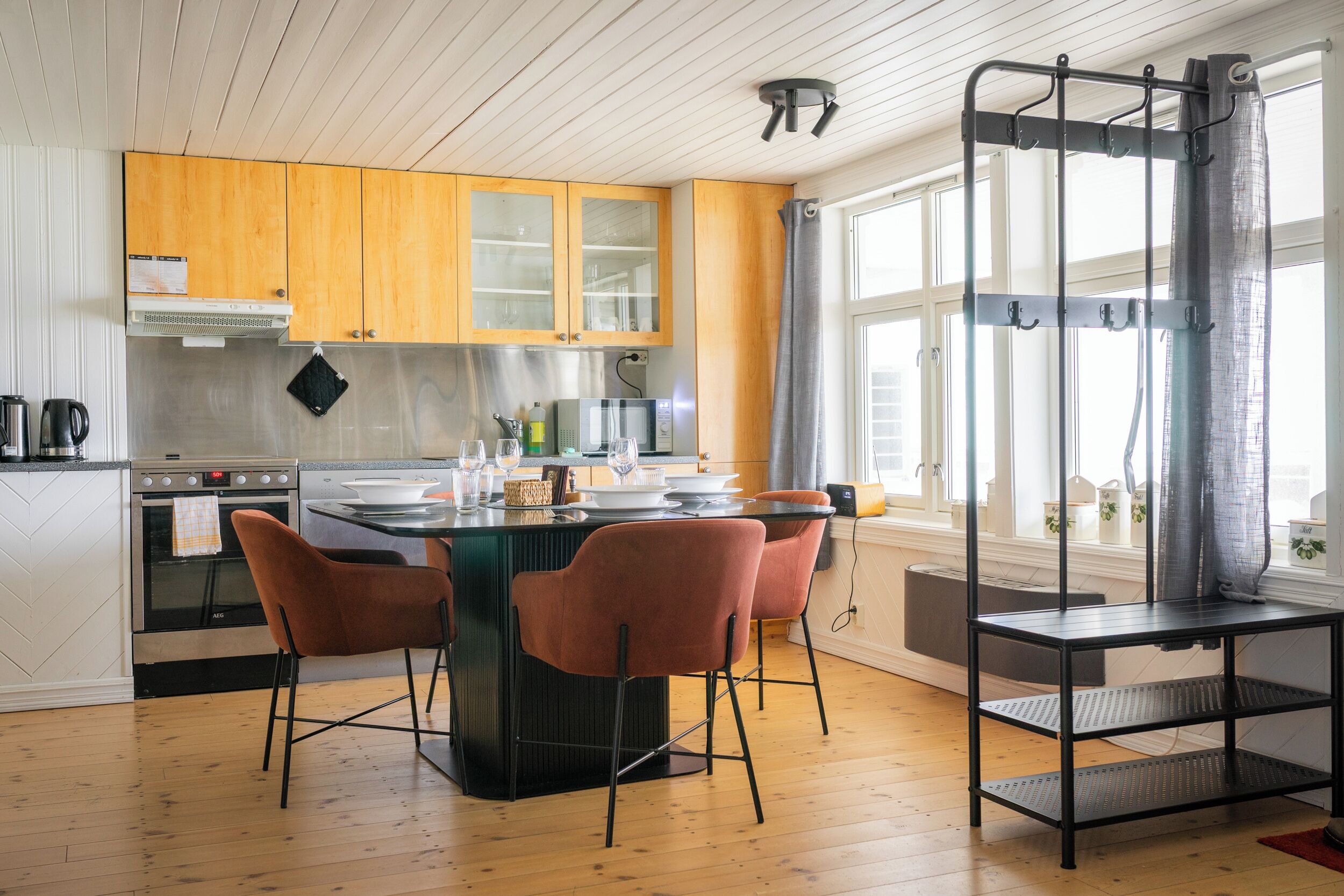 Una cocina moderna con armarios de madera, una isla central con sillas, un estante de metal negro y una ventana con cortinas.