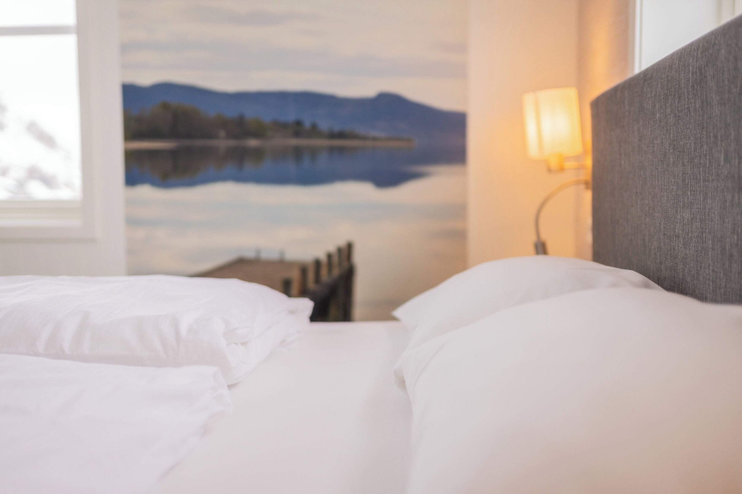 Habitación de hotel con cama, mesita de noche, lámpara y un cuadro en la pared que muestra un lago y montañas.