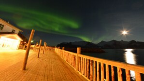 Una terraza de madera con barandilla, un edificio con entrada iluminada y montañas al fondo.
