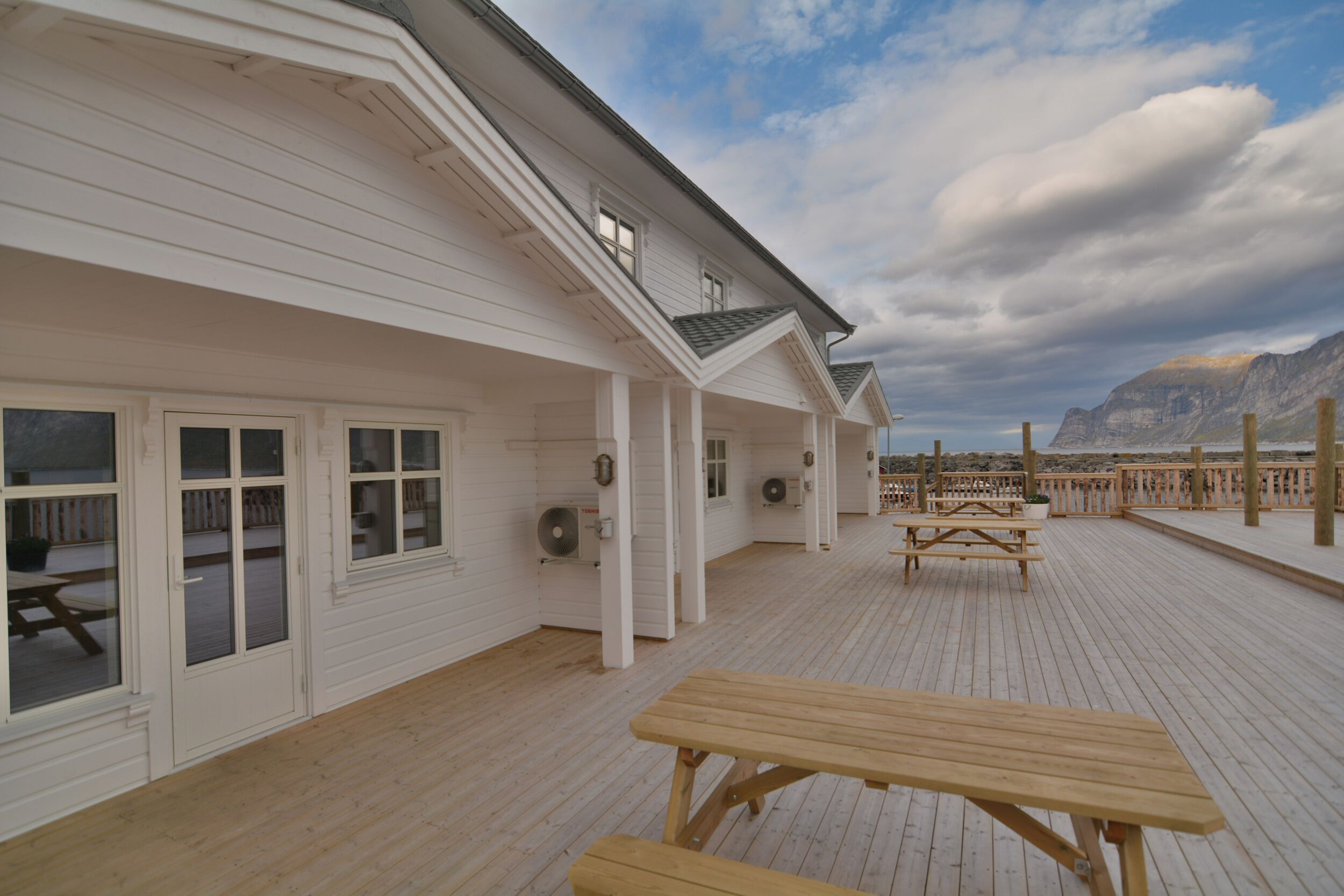 A white building with a wooden deck, picnic tables, and a view of a mountainous landscape.