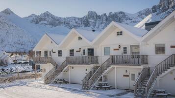 A row of white cabins with wooden balconies and stairs, set against a snowy mountain backdrop.