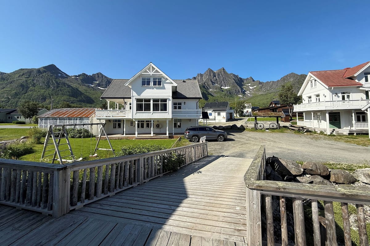 Une terrasse en bois mène à un bâtiment blanc doté de nombreuses fenêtres et d’un balcon, le tout étant entouré de montagnes.