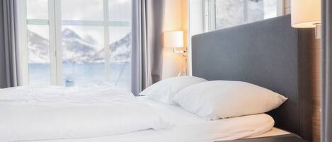 A modern hotel room with a large bed, a bedside table, a lamp, and a view of mountains through large windows.