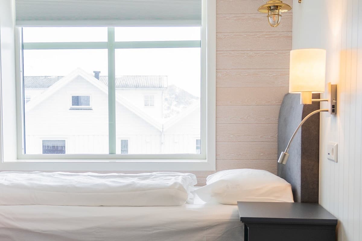 Un lit bien fait, avec du linge de lit blanc, une table de chevet avec une lampe, et une vue sur les bâtiments par une grande fenêtre.