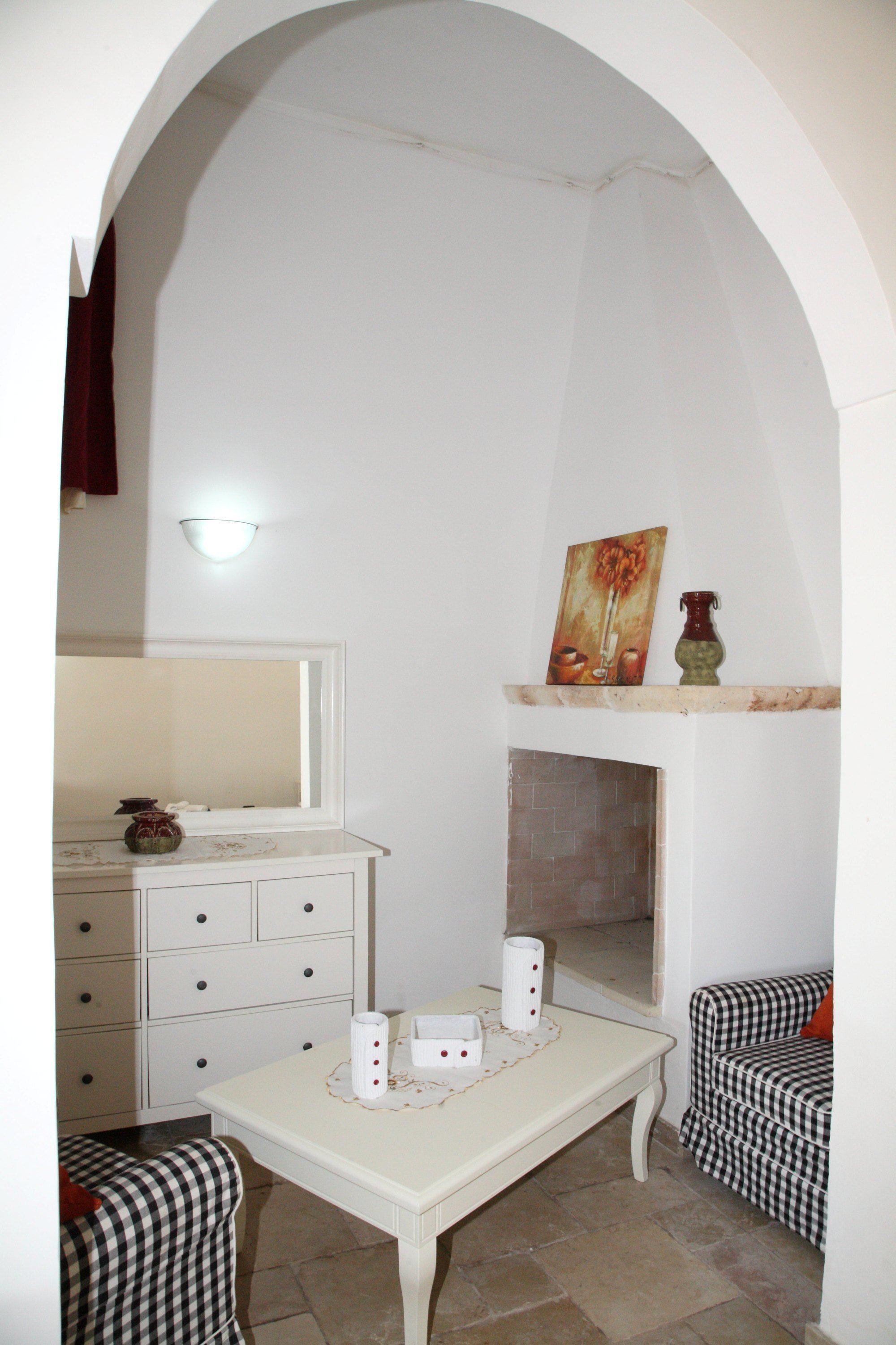 Een kamer met een witte boogdoorgang, een witte tafel met een vaas en kaarsen, een geruit stoel, en een open haard met een schilderij erboven.