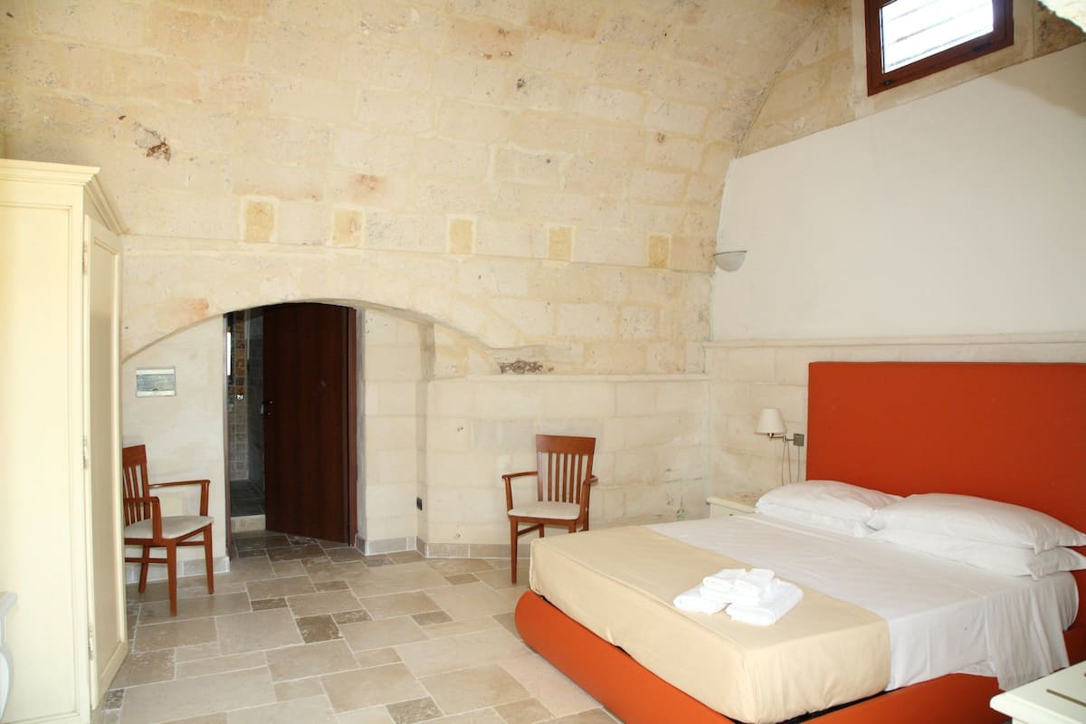 Une chambre avec un mur en pierre, un lit doté d’une tête de lit orange, une chaise en bois et une petite table.