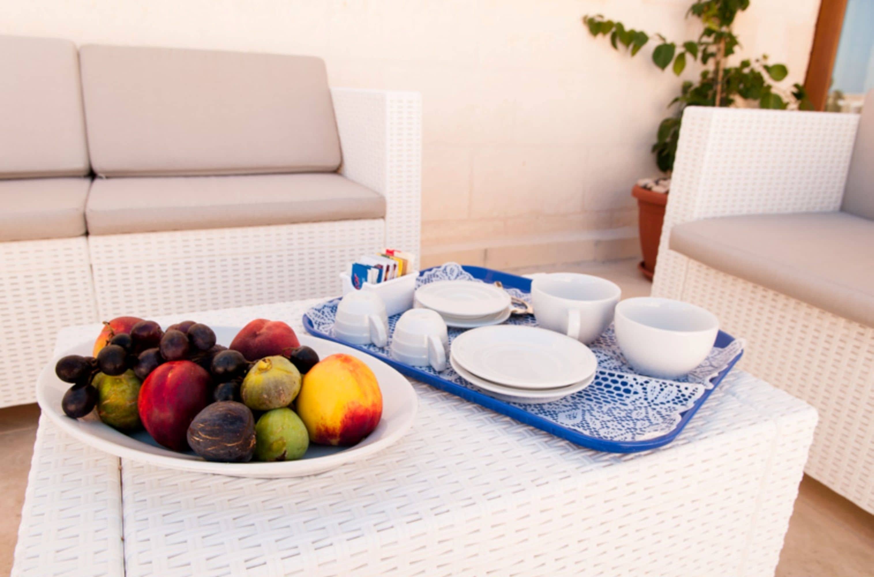 A wicker sofa, a tray with fruits and dishes, and a potted plant in a sunny outdoor setting.